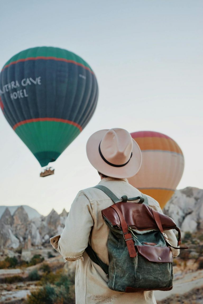 Traveler with backpack watching hot air balloons soar over Cappadocia's unique landscape at sunrise.
